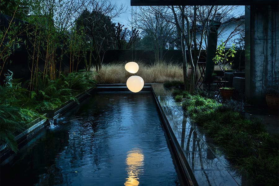 Giardino con le lampade da terra Foscarini Gregg a sasso di vetro accese lungo una vasca d'acqua, al tramonto: la stessa scena al buio e con la luce accesa