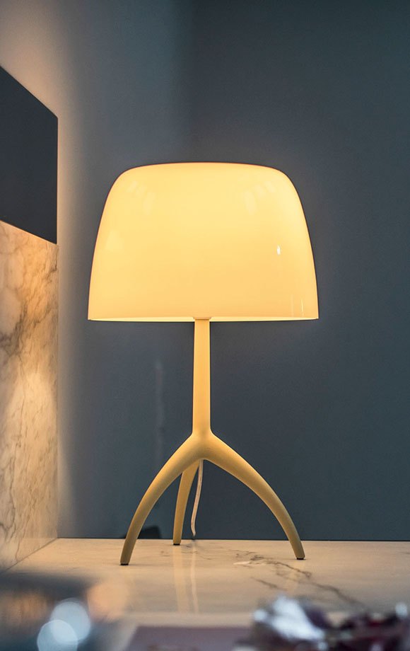 Lampada da tavolo Foscarini Lumiere con il diffusore di vetro acceso accanto a una finestra