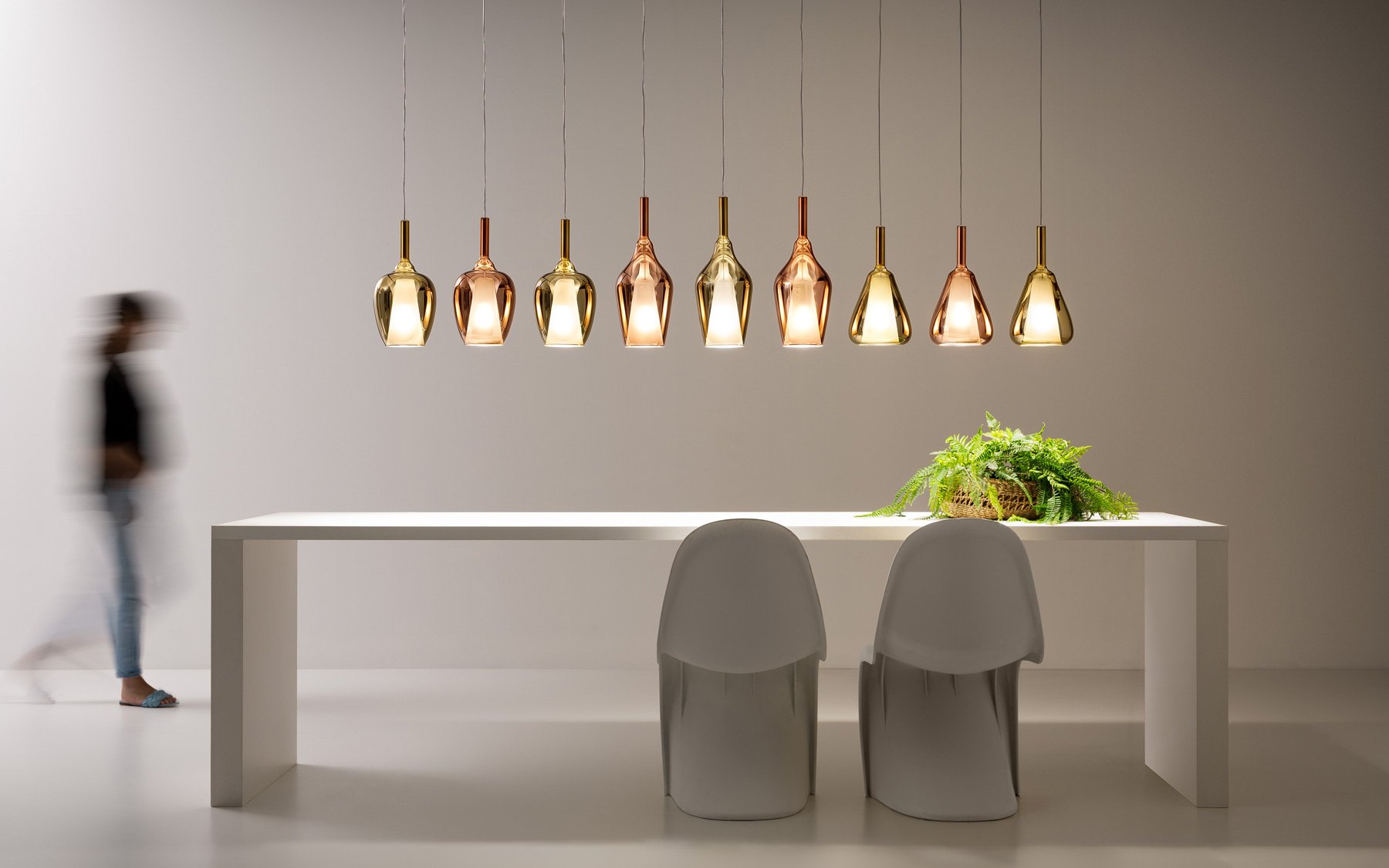 Sala da pranzo con le sospensioni Gea Luce in vetro ambra in fila sopra il tavolo bianco: la stessa scena al buio e con la luce accesa