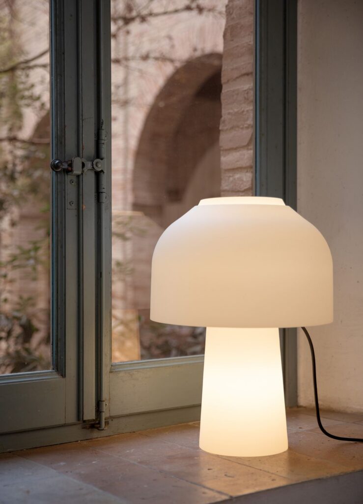 Lampada da tavolo Karman Lilly in vetro bianco davanti a una finestra
