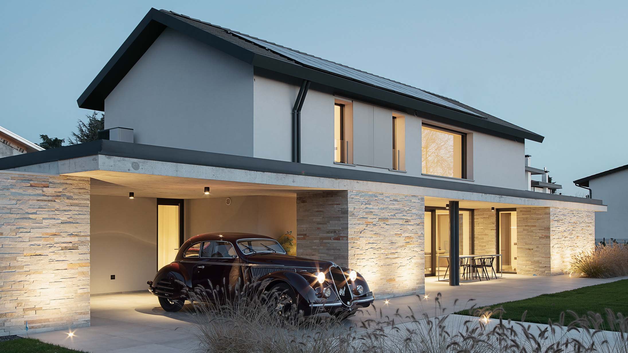 Villa moderna con la facciata illuminata di sera e un'auto d'epoca davanti: la stessa scena al buio e con la luce accesa
