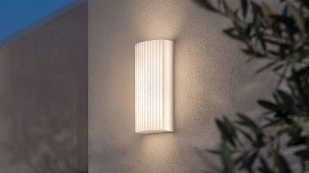 Applique da esterno Lombardo Glow con il diffusore cannettato, accanto a un ulivo